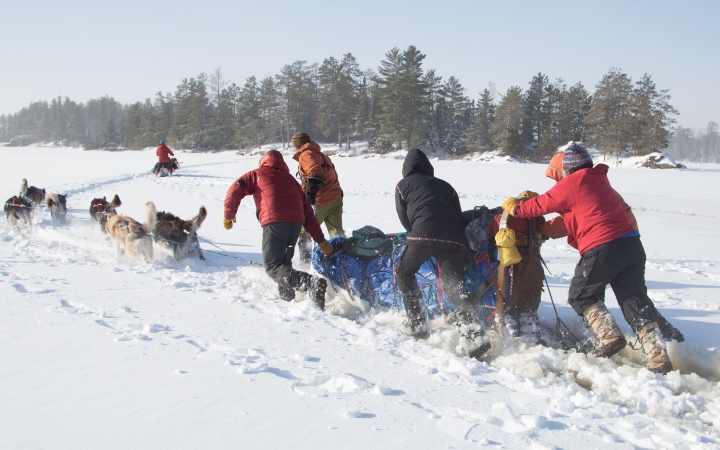 Four people push a sled through slushy conditions behind a team of dogs. There is another team ahead of them. 